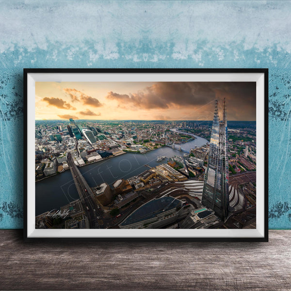The image is a framed aerial view of a cityscape, showcasing a mix of tall buildings, roads, and a river. The sky is filled with clouds, creating a dramatic backdrop for the city's skyline. The image is framed and displayed on a wooden surface, likely a wall or shelf.
