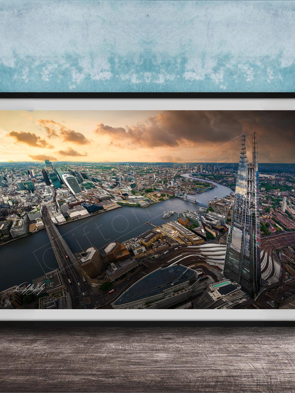 The image is a framed aerial view of a cityscape, showcasing a mix of tall buildings, roads, and a river. The sky is filled with clouds, creating a dramatic backdrop for the city's skyline. The image is framed and displayed on a wooden surface, likely a wall or shelf.