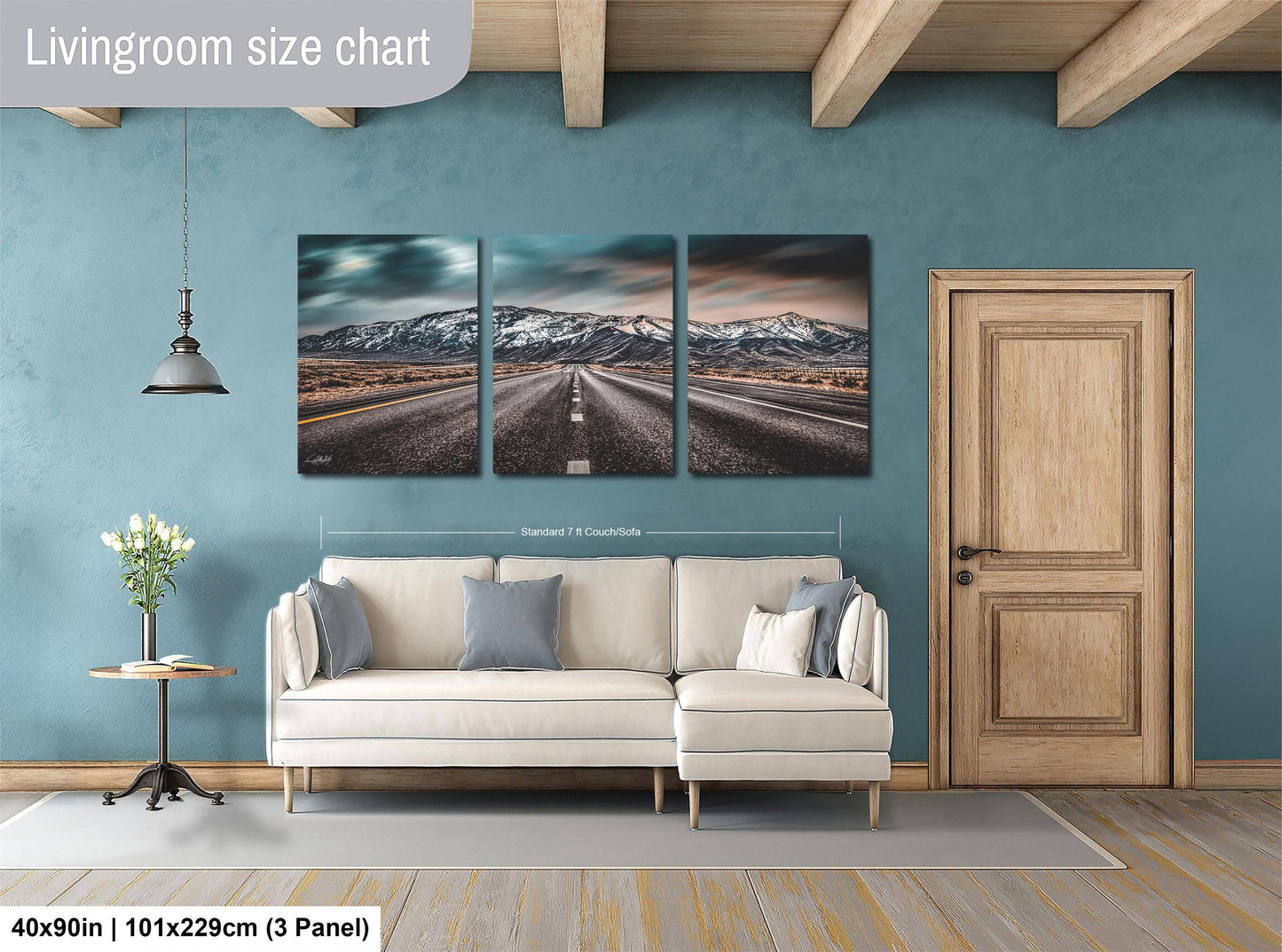 The image is a living room with a large white sofa, a wooden door, and a wall with a triptych of landscape paintings depicting a road leading to a mountain range.