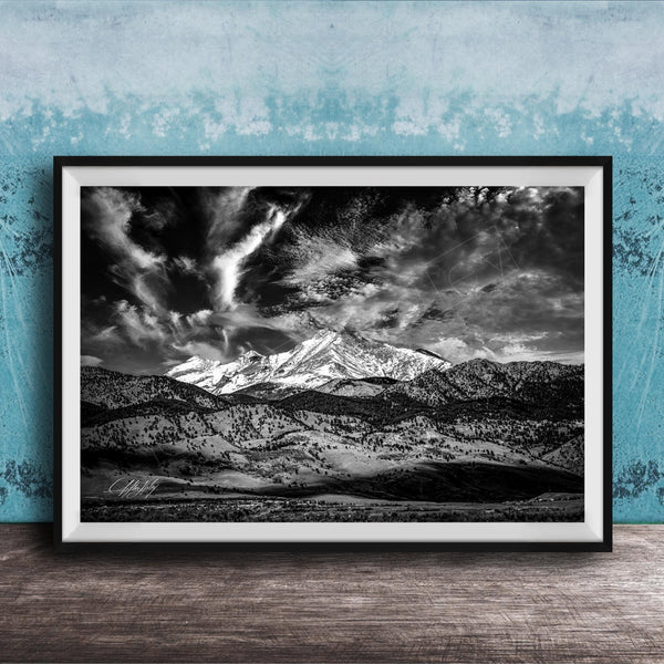 A black and white photograph of a mountainous landscape with snow-capped peaks and dramatic clouds in the sky, framed and displayed on a wooden surface against a blue textured wall.