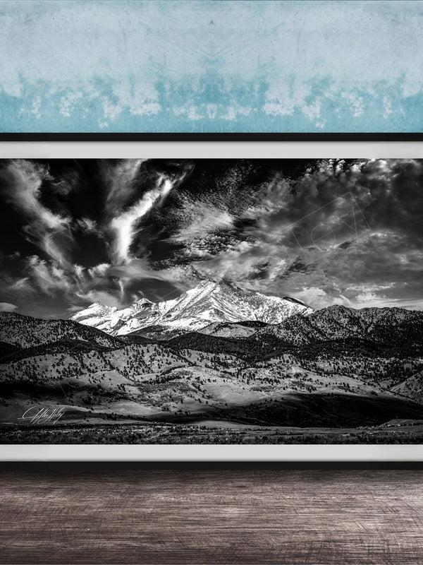A black and white photograph of a mountainous landscape with snow-capped peaks and dramatic clouds in the sky, framed and displayed on a wooden surface against a blue textured wall.