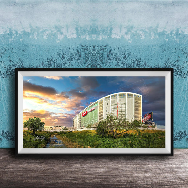 A framed photograph of a large building with a curved roof, surrounded by trees and a river, with a colorful sunset sky in the background.