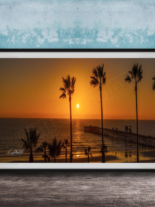 A framed photograph of a serene beach scene at sunset, with palm trees silhouetted against the warm orange sky.