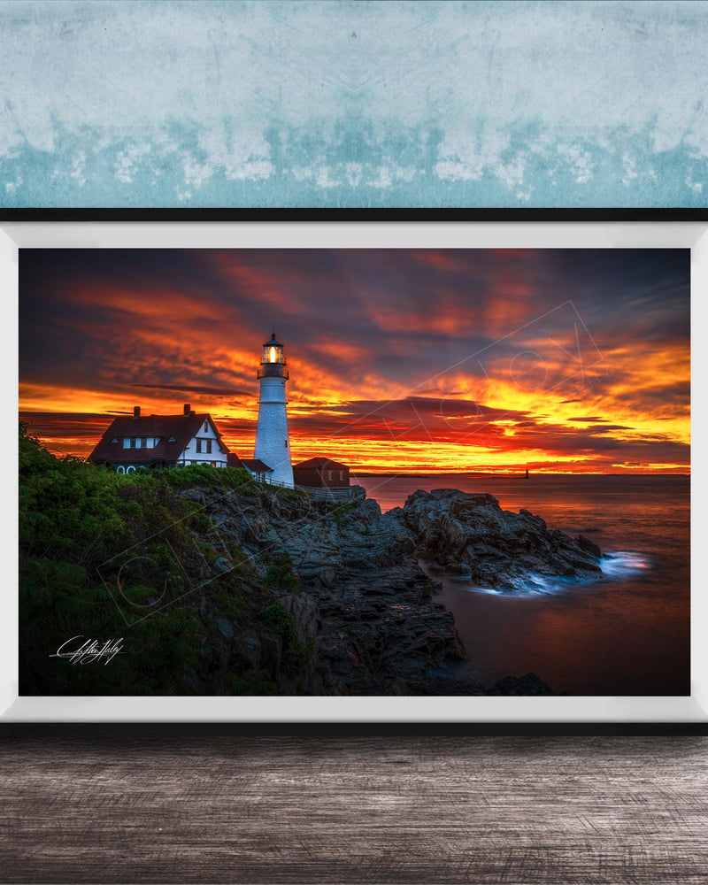 A framed photograph depicting a lighthouse on a rocky cliff at sunset, with the sky filled with vibrant orange and red hues.