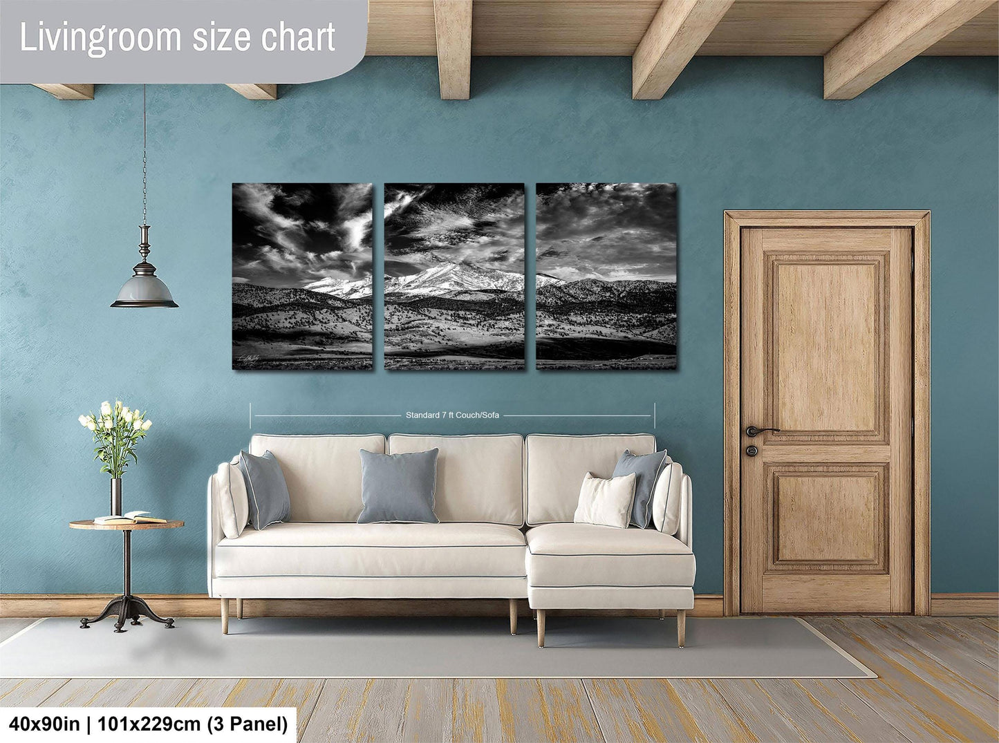 The image is a living room with a large, three-panel landscape painting hanging on the wall. The painting depicts a mountain range with a cloudy sky. The room has a white sofa and a wooden door, and there is a vase of flowers on a table. The painting is approximately 40 inches wide and 101.29 cm tall, with each panel measuring approximately 21 inches wide and 29.7 cm tall.