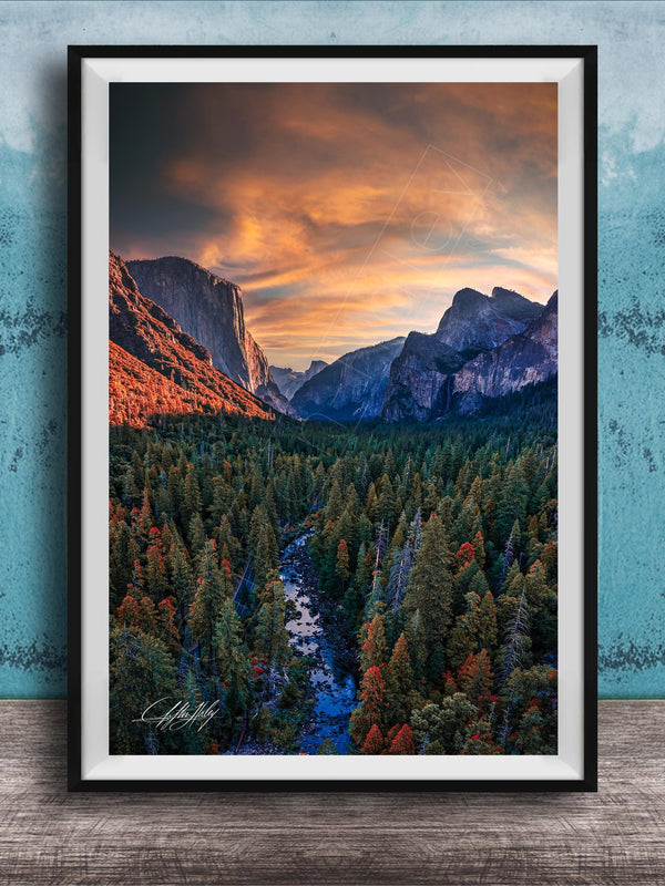 The image is a framed photograph of a mountainous landscape with a river running through it. The mountains are covered in trees, and the sky is filled with clouds, creating a dramatic and colorful scene.