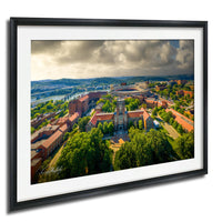 The image is a framed aerial view of a city with a mix of buildings, trees, and a river. The sky is cloudy, and the overall scene has a somewhat gloomy atmosphere.
