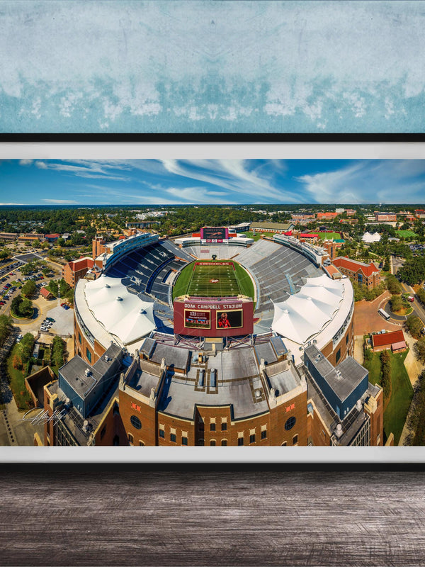 The image is an aerial view of a large sports stadium with a football field, surrounded by buildings and a blue sky.