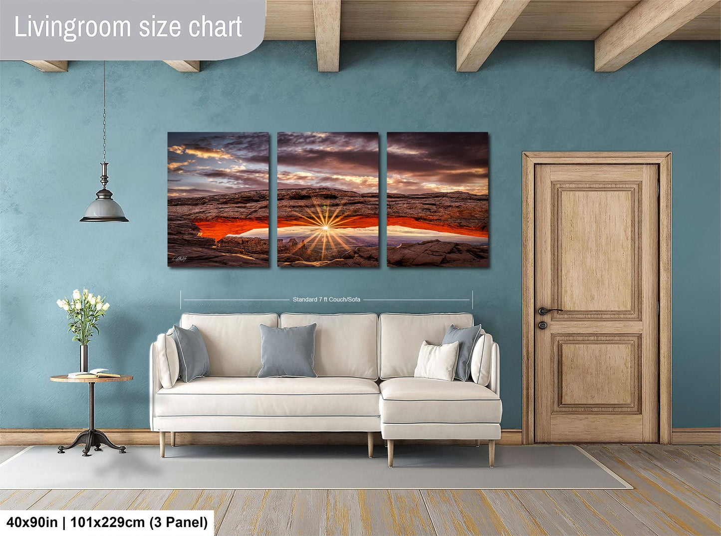 The image is a living room with a large, three-panel landscape painting hanging on the wall. The painting depicts a scenic sunset over a rocky landscape. The room has a white sofa and a wooden door, creating a cozy and inviting atmosphere.
