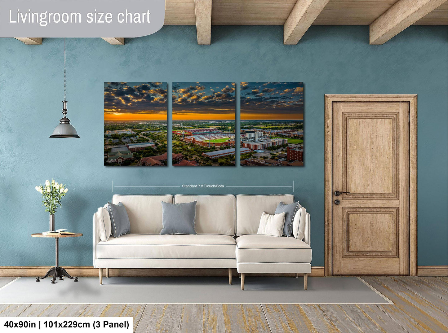 The image is a living room with a large, three-panel landscape painting hanging on the wall. The painting depicts a cityscape with buildings and a sunset in the background. The room has a white sofa and a wooden door, and there is a vase of flowers on a table. The painting is approximately 40 inches wide and 101.29 cm in height, with each panel measuring approximately 3 feet in width.