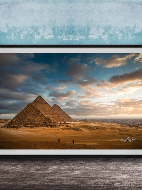 two large sandstone pyramids against a cloudy sky, with a few people walking in the foreground.