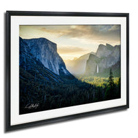 a breathtaking view of the majestic Yosemite Falls, with its powerful waterfall cascading down the towering granite cliffs. The sky above is a beautiful canvas of orange and yellow hues, creating a stunning backdrop for the natural landscape. The frame is black, which contrasts with the vibrant colors of the scene, making it stand out even more.