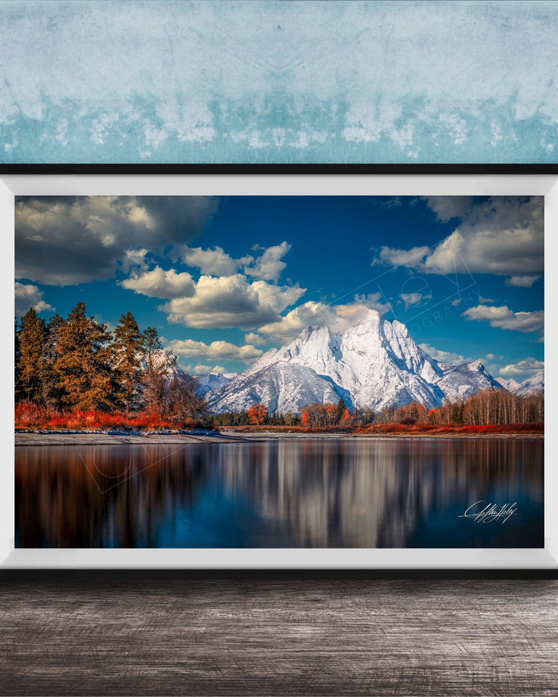 A framed photograph depicting a serene mountain landscape with a calm lake reflecting the surrounding scenery, including trees and mountains in the background.