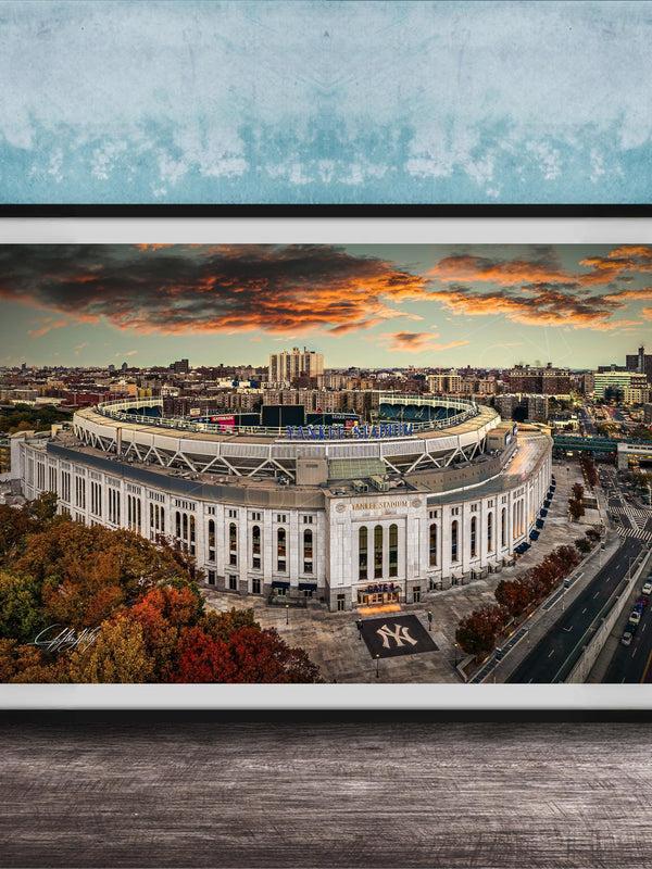 The image is a framed photograph of the New York Yankees stadium, which is a large, circular stadium located in the city of New York. The photograph captures the stadium's distinctive architecture and surrounding cityscape, with a dramatic sky in the background.