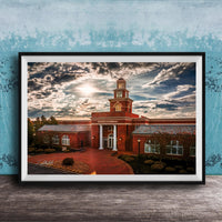 A framed photograph of a red brick building with a tower, set against a cloudy sky at sunset.