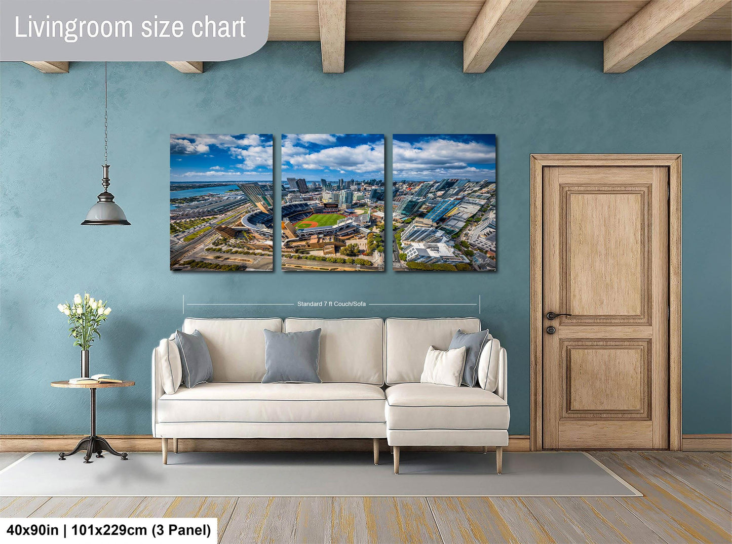 The image is a living room with a large wall-mounted painting depicting a cityscape. The painting is divided into three panels, each showing different parts of the city. The room has a white sofa, a wooden door, and a small table with a vase of flowers. The painting is approximately 40 inches wide and 101.29 cm tall, with a 3-panel arrangement.