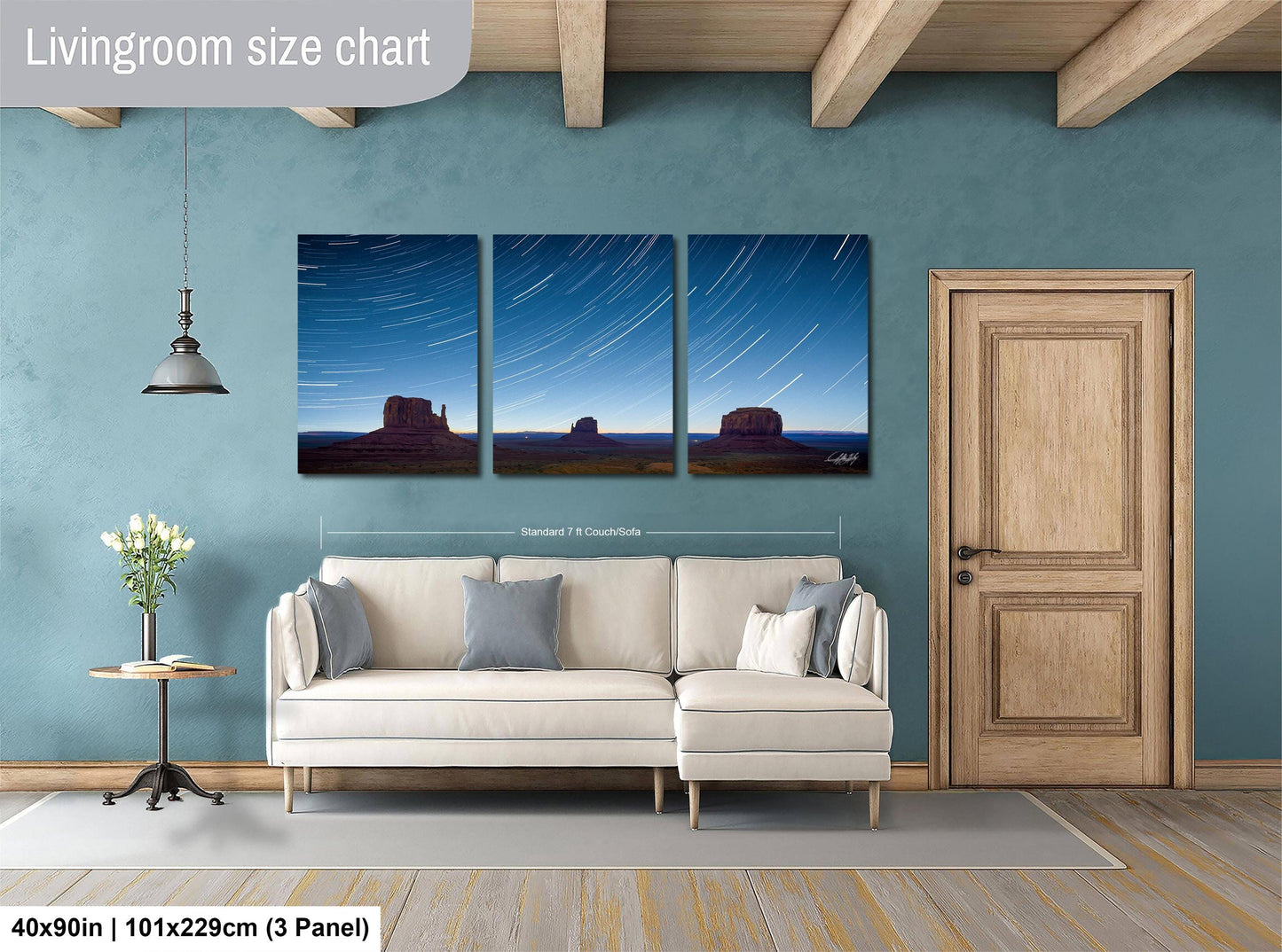 The image is a living room with a large wall art depicting a night sky with stars and a desert landscape. The wall art is divided into three panels, each showing a different part of the night sky and desert scene. The living room has a white sofa, a wooden door, and a small table with a vase of flowers. The wall art is approximately 40 inches wide and 101.29 cm tall, with a 3-panel layout.