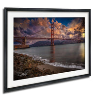 a picturesque scene of the Golden Gate Bridge in San Francisco, California, with its iconic red towers and suspension cables spanning the bay. The bridge is set against a dramatic sky filled with clouds and vibrant colors, creating a stunning backdrop for the serene waters of the bay. The photograph is framed and displayed on a wall, showcasing the beauty of this iconic landmark.