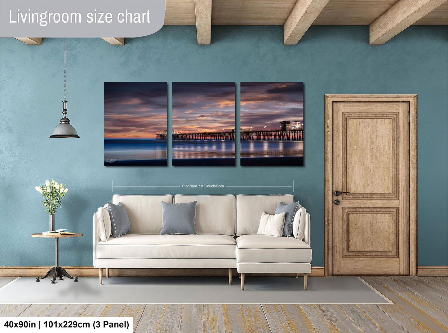 The image is a living room with a large wall art piece depicting a serene sunset over a body of water, featuring a pier and a bridge. The living room has a white sofa and a wooden door, and the wall art is approximately 40 inches wide and 101.29 cm in height.