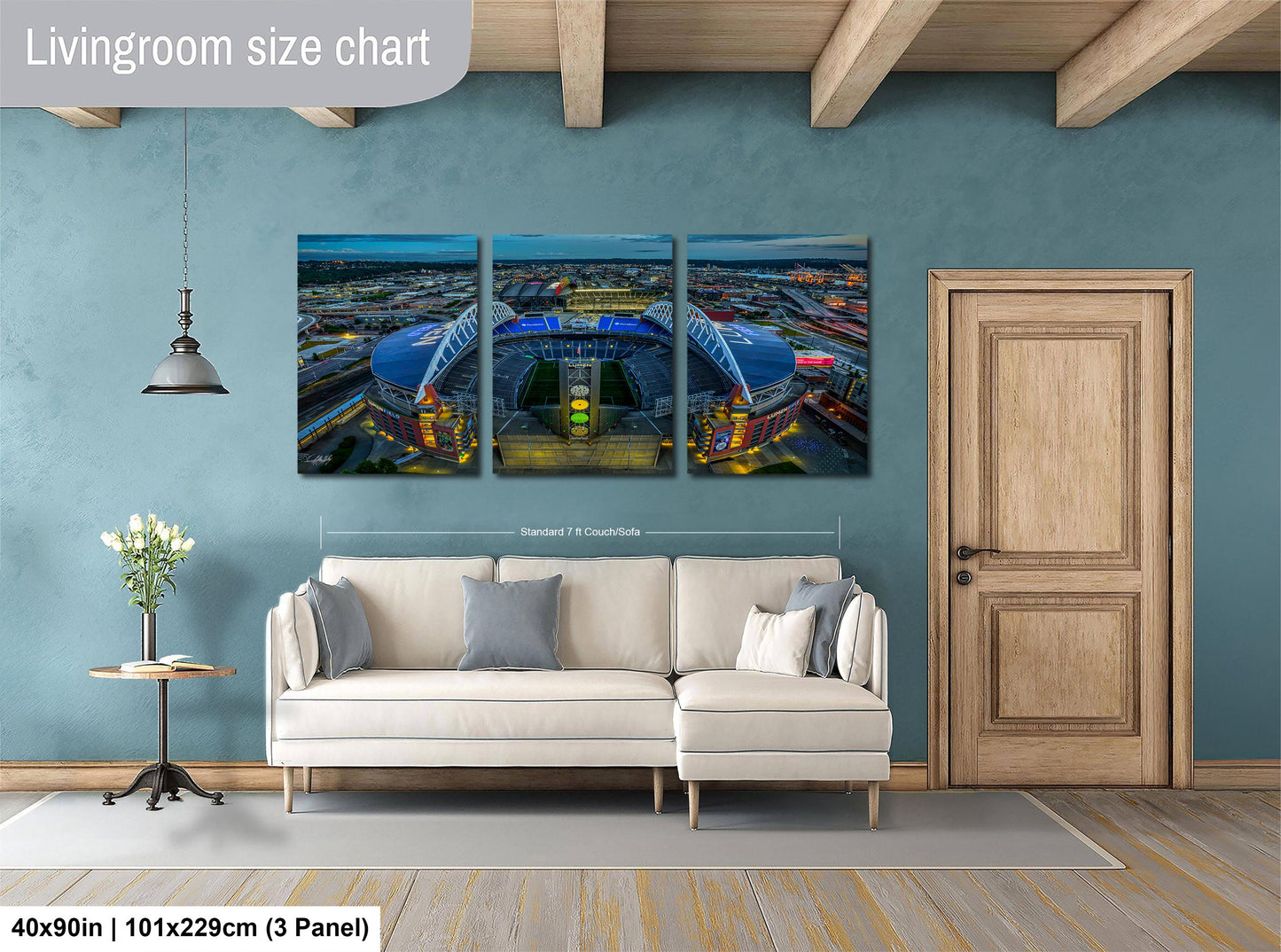 The image is a living room with a large wall art depicting a city skyline at night. The wall art is divided into three panels, with the largest panel covering the entire wall. The room features a white sofa, a wooden door, and a small table with a vase of flowers. The wall art is approximately 40 inches wide and 101.29 cm tall, with a 3-panel arrangement.