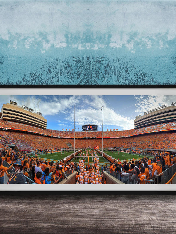 A framed photograph of a crowded football stadium with a large crowd of spectators, a large scoreboard, and a large field.