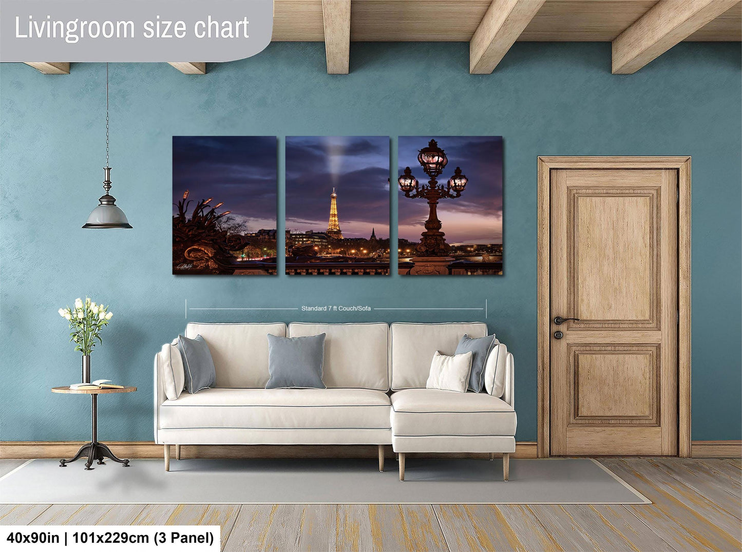 The image is a living room with a white sofa and a wooden door. Above the sofa, there is a large wall art collage depicting a city skyline at night, including a lit lamp post and a tall tower. The collage is divided into three panels, each showing different parts of the city skyline. The wall art is framed by a wooden border and is approximately 40 inches wide and 101.29 cm in height.