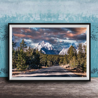A framed photograph depicting a scenic mountain landscape with a winding road surrounded by pine trees and snow-capped peaks under a dramatic cloudy sky.