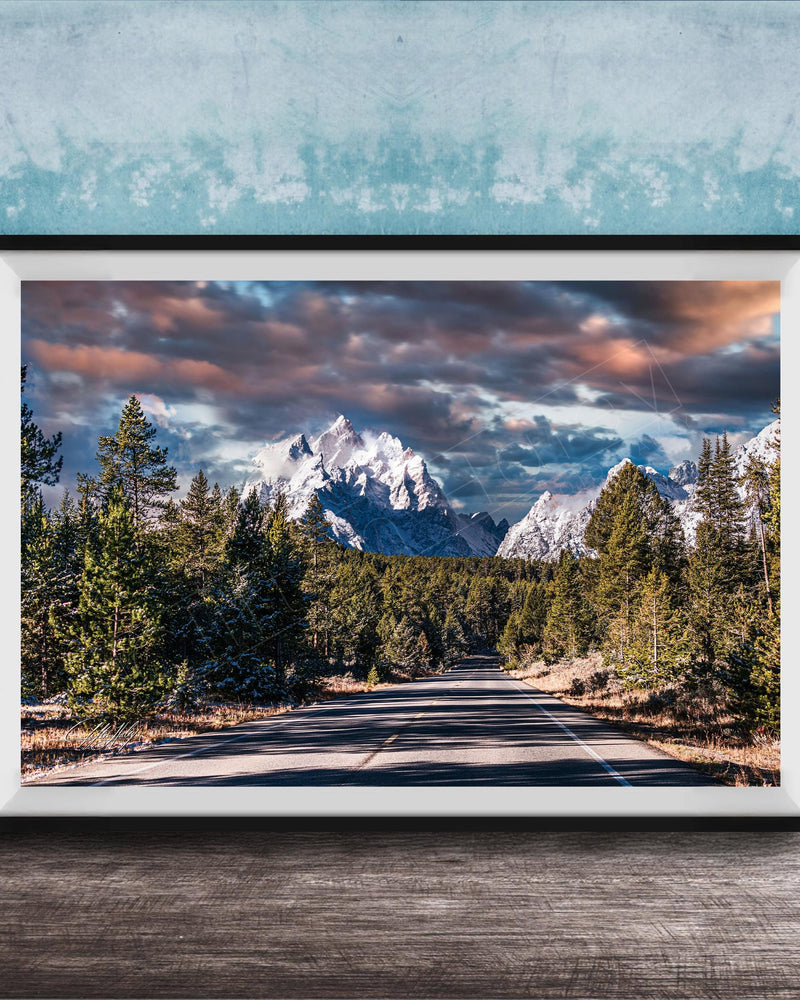 A framed photograph depicting a scenic mountain landscape with a winding road surrounded by pine trees and snow-capped peaks under a dramatic cloudy sky.