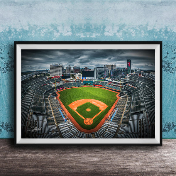 A framed photograph of a baseball stadium with a large field, surrounded by buildings and a cloudy sky.