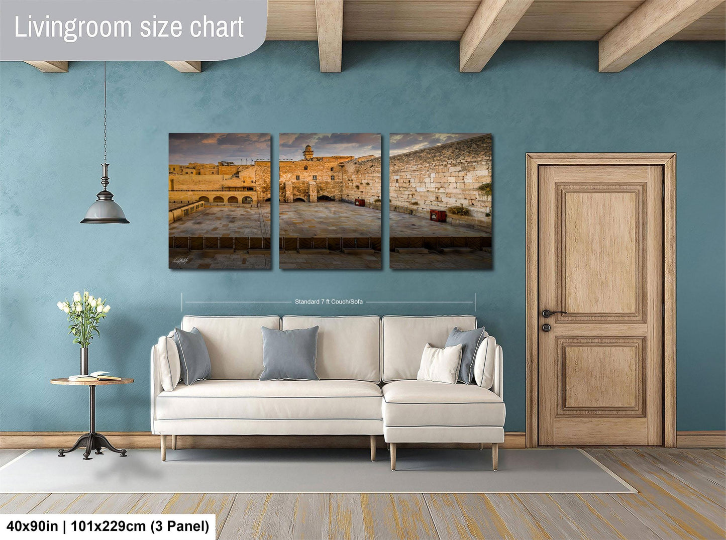 The image is a living room with a white sofa and a wooden door. Above the sofa, there is a triptych of pictures depicting a cityscape, including a building and a street. The wall behind the sofa has a blue color, and there is a lamp hanging from the ceiling. The image is described as a living room size chart, indicating that it is a visual representation of the dimensions of the living room.