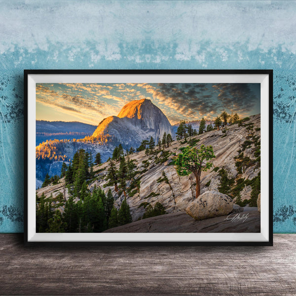 The image is a framed photograph depicting a mountainous landscape with a large mountain peak in the background, surrounded by trees and rocky terrain. The sky is filled with warm, golden light, creating a picturesque and serene atmosphere.