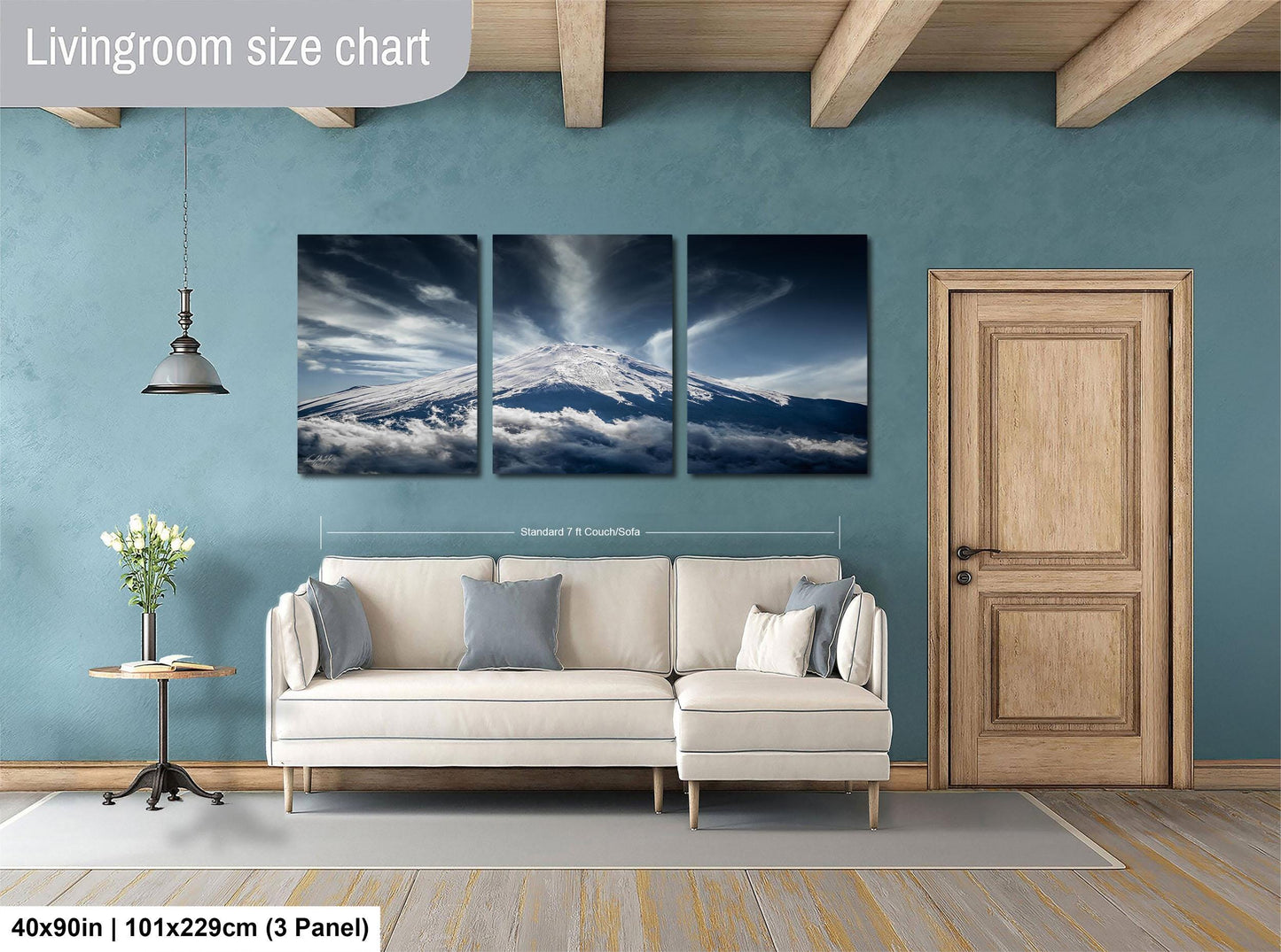 The image is a living room with a large wall art depicting a mountain landscape with clouds. The wall art is divided into three panels, and the living room has a white sofa, a wooden door, and a small table with a vase of flowers.
