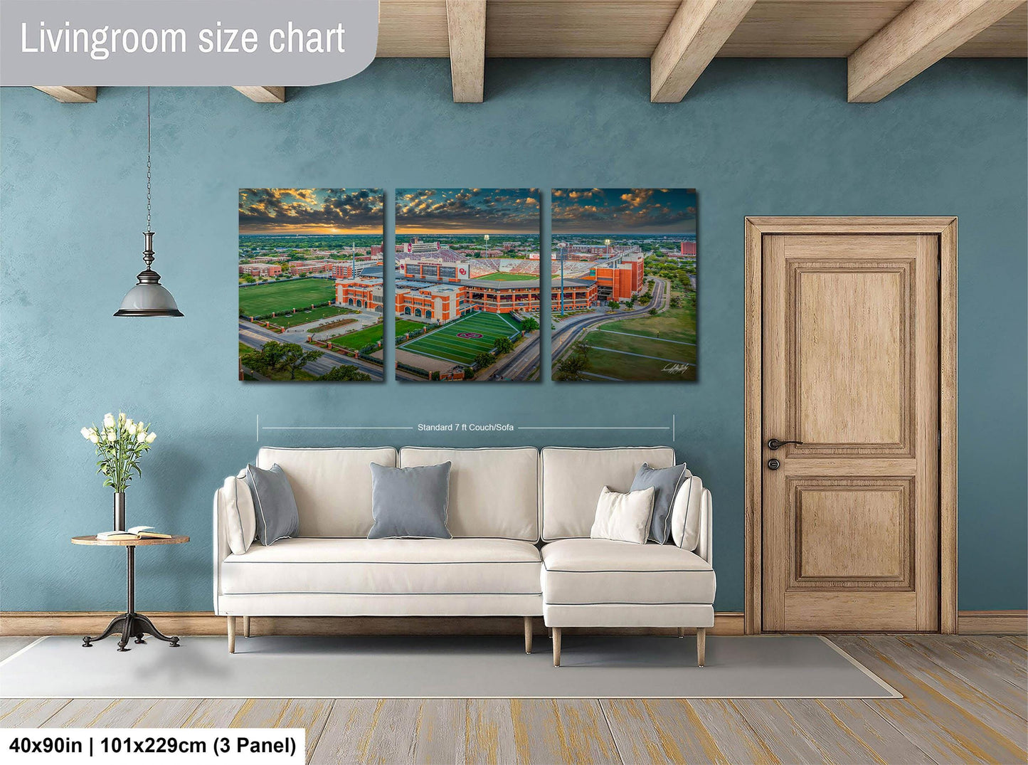 The image is a living room with a large, three-panel wall art depicting a cityscape. The wall art is mounted above a white sofa, and there is a wooden door and a small table with a vase of flowers nearby. The wall art is approximately 40 inches wide and 101.29 cm in height, with each panel measuring approximately 3 feet in width.