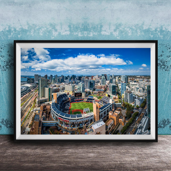 An aerial view of a large city with a baseball stadium in the center, surrounded by tall buildings and a blue sky with clouds.