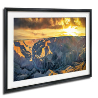 The image is a framed painting depicting a breathtaking view of the Grand Canyon at sunset. The painting showcases the vast, rugged landscape with dramatic cliffs and deep valleys, illuminated by the warm, golden light of the setting sun. The colors in the painting are vibrant, with the orange and yellow hues of the sunset contrasting beautifully with the cool blues and greens of the canyon. The painting is framed in a black frame, which enhances the visual impact of the scene.