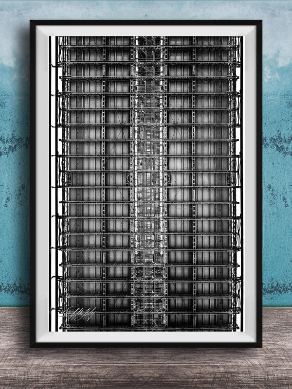 The image is a black and white photograph of a tall, narrow building with a grid-like pattern of windows and steel beams. The photograph is framed and displayed on a wooden surface against a blue wall.