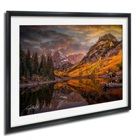 a serene mountain landscape with a lake reflecting the vibrant colors of the autumn foliage. The mountains are adorned with a mix of green and orange hues, creating a picturesque scene. The sky above is filled with clouds, adding to the dramatic atmosphere of the landscape. In the foreground, there are two moose standing near the lake, adding a touch of wildlife to the scene. The image is framed and appears to be a painting or photograph, capturing the beauty of nature.