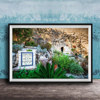 A framed photograph depicting a stone wall with a sign that reads "JESUS CHRIST" and "SON OF GOD". The wall is surrounded by lush greenery, including plants and trees.