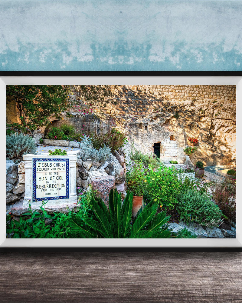 A framed photograph depicting a stone wall with a sign that reads "JESUS CHRIST" and "SON OF GOD". The wall is surrounded by lush greenery, including plants and trees.