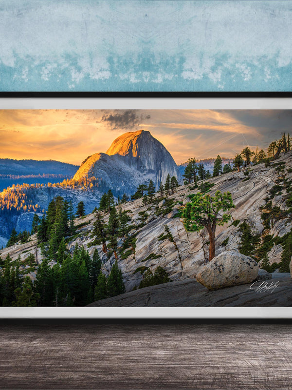 A framed photograph depicting a mountainous landscape with a large, snow-capped peak in the distance, surrounded by trees and rocky terrain. The sky is filled with warm, golden light, creating a serene and picturesque scene.