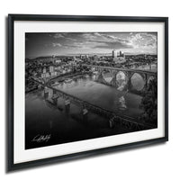 A black and white aerial view of a city with a large bridge spanning a river, surrounded by buildings and hills.