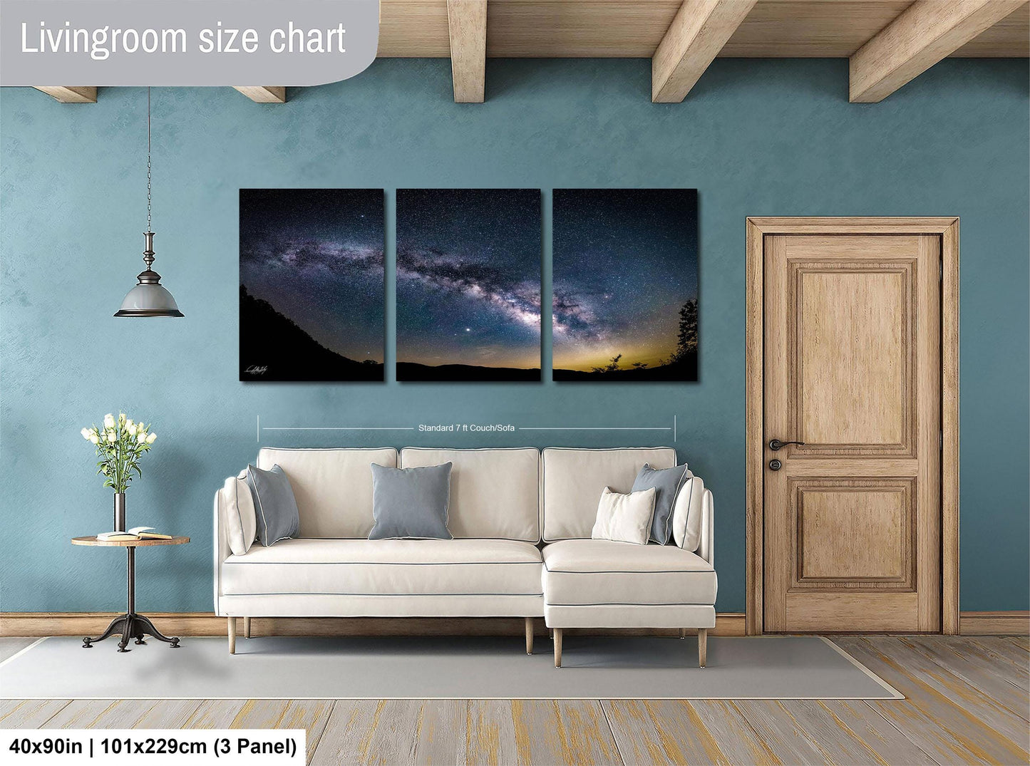 The image is a living room with a large wall art depicting a night sky with a galaxy. The wall art is divided into three panels, each showing a different part of the night sky. The living room has a white sofa, a wooden door, and a small table with a vase of flowers. The wall art is approximately 40 inches wide and 101.29 cm tall, with a 3-panel layout.