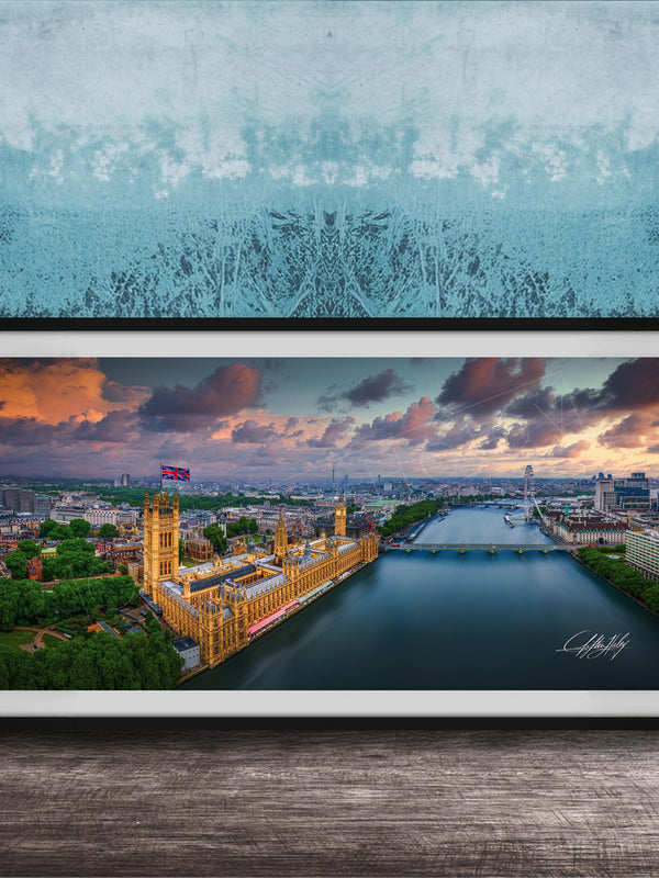 The image is a framed photograph of the Palace of Westminster, also known as the Houses of Parliament, in London, England. The photograph captures the iconic skyline of the city, with the River Thames flowing in the foreground. The sky is filled with vibrant colors, suggesting a beautiful sunset or sunrise. The photograph is framed and displayed on a wooden surface, creating a visually appealing and artistic composition.