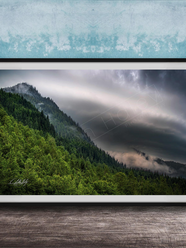 The image is a framed photograph of a mountainous landscape with a forested area and a cloudy sky.
