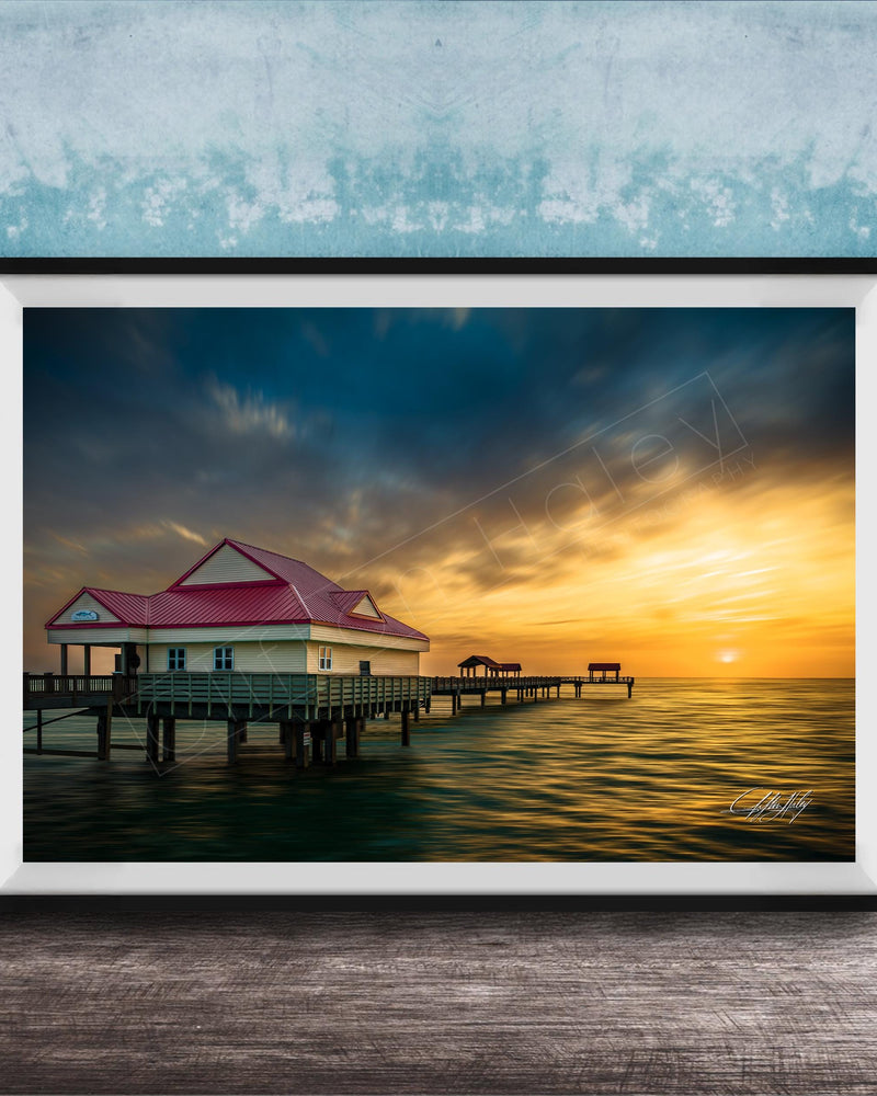 A framed photograph depicting a serene pier scene at sunset, with a red-roofed building and a wooden pier extending into the calm waters.