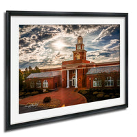 A framed photograph of a brick building with a prominent tower, set against a cloudy sky with the sun peeking through.