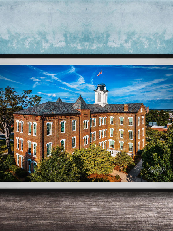 A framed photograph of a large brick building with a clock tower, surrounded by trees and a blue sky with clouds.