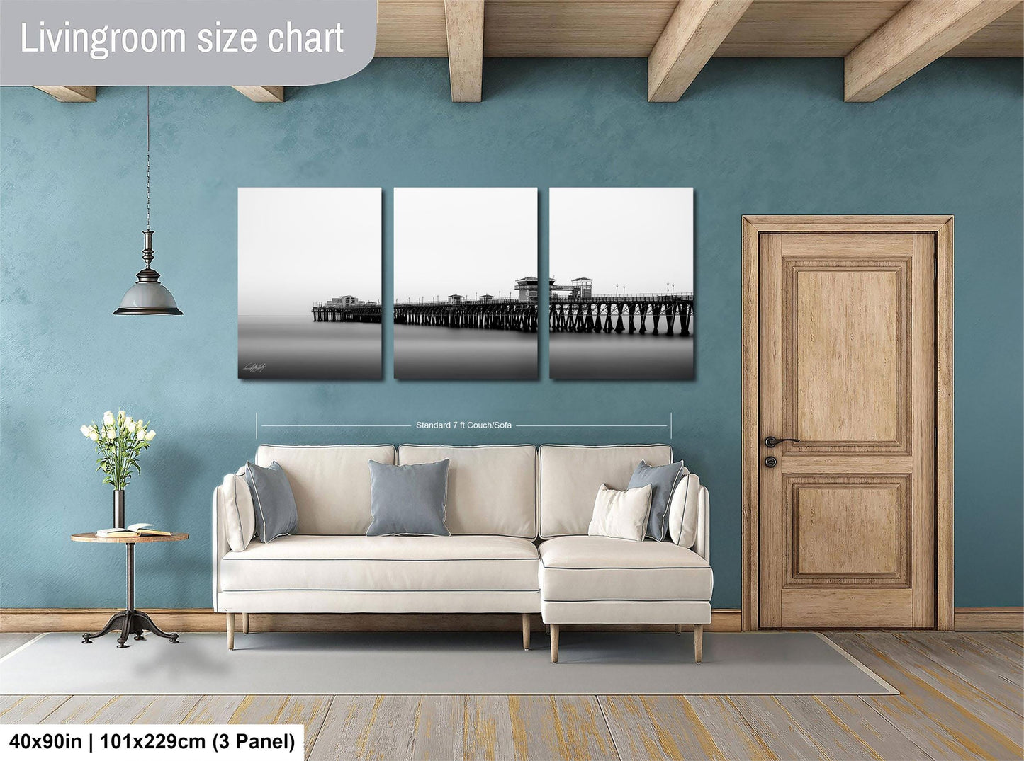 The image is a living room with a large white sofa, a wooden door, and a wall with a large black and white photograph of a pier and a bridge.