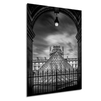 A black and white photograph of the Louvre Museum in Paris, France, with the iconic glass pyramid structure of the museum prominently featured. The photograph is captured from a low angle, emphasizing the grandeur and architectural details of the building.