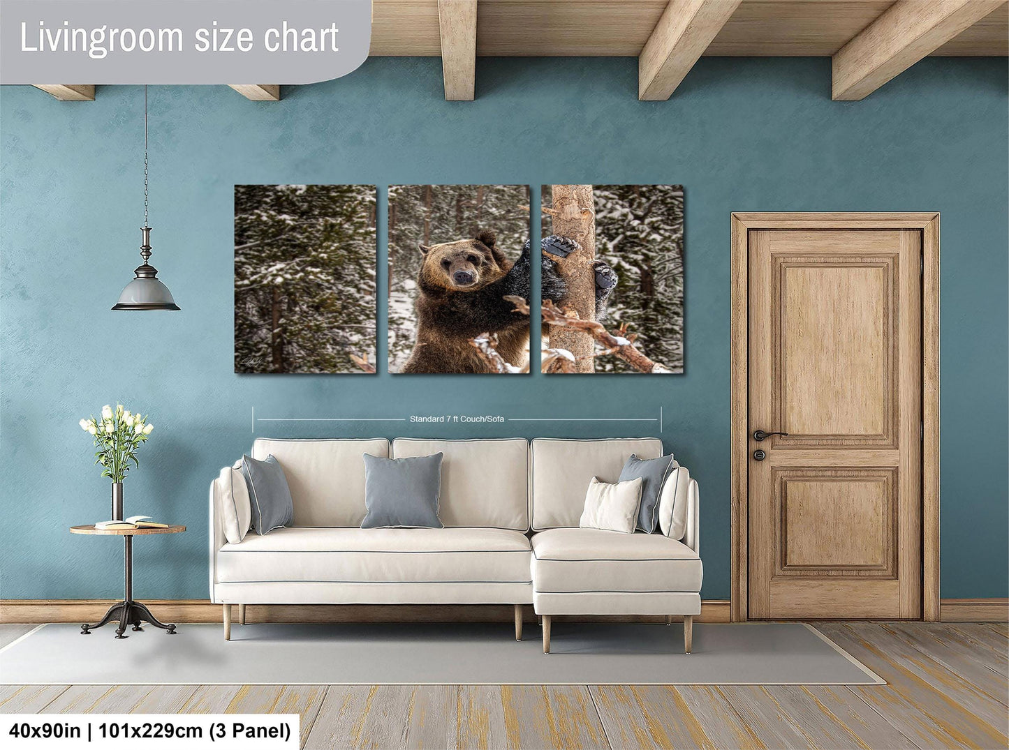 The image is a living room with a large wall art piece depicting a brown bear climbing a tree. The wall art is divided into three panels, with the largest panel showing the bear in the center. The room has a white sofa, a wooden door, and a small table with a vase of flowers. The wall art is approximately 40 inches wide and 101.29 cm tall, with a 3-panel layout.